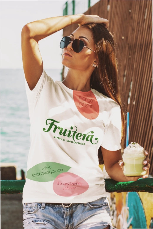 A young woman enjoys a smoothie on the beach, wearing a Fruitera t-shirt, with waves and sand in the background.